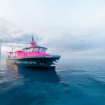 The pink vessel floats on a calm ocean surface, with wind turbines visible in the background, symbolizing the integration of renewable energy and cutting-edge marine technology.